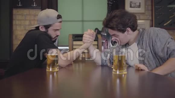 两个人坐在酒吧的一张桌子旁从事手臂摔跤一个人赢了另一个人输了两个人都喝啤酒伙计们视频的预览图