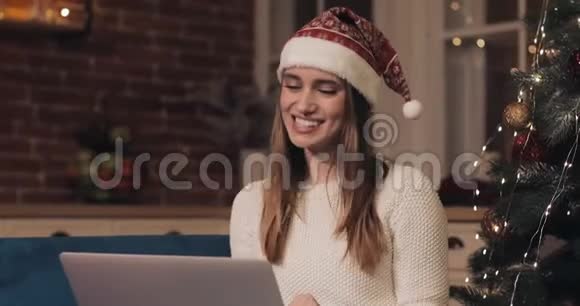 一位戴着圣诞帽笑容满面的白种人女士坐在圣诞树旁舒适的家中的沙发上打着视频电话视频的预览图