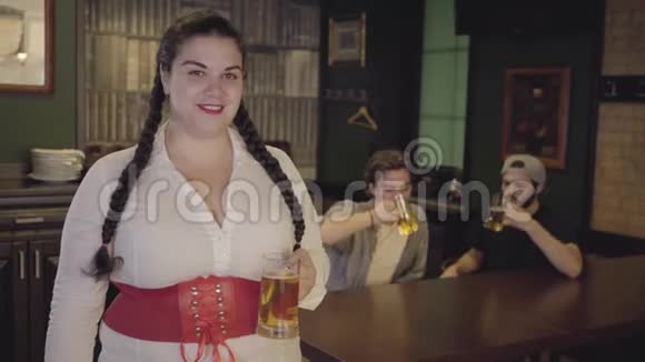 一个丰满的女人梳着白衬衫的辫子胸衣拿着啤酒杯看着两个男人在酒吧里喝酒视频的预览图