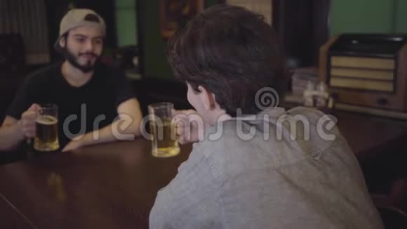 两个人在酒吧里坐在一张桌子旁喝着啤酒喝着叮当作响的眼镜一起喝啤酒玩得开心休闲视频的预览图