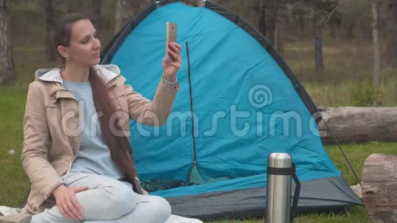 一个留着长发的年轻女孩坐在森林里的一个蓝色帐篷附近在电话里自拍视频的预览图