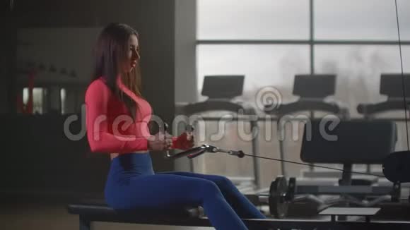 一个坐着的女人在健身房的大窗户和跑步机的背景下拉动模拟器的重量视频的预览图