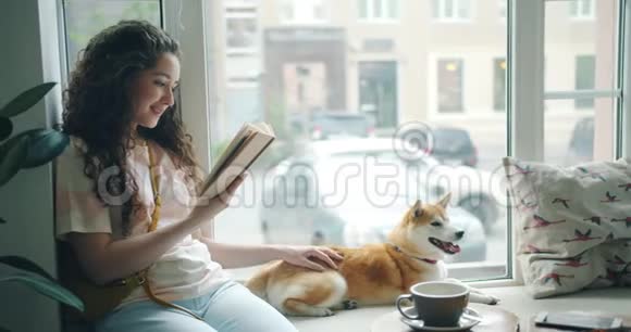 漂亮的年轻女士在窗台的咖啡馆里读小说抚摸宠物狗视频的预览图