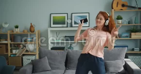 美丽的女士在家跳舞戴着耳机手持智能手机唱歌视频的预览图