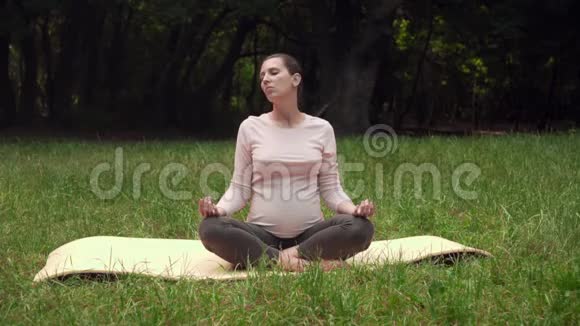 孕妇在公园里地毯上做瑜伽坐在莲花的位置热身颈部和头部视频的预览图