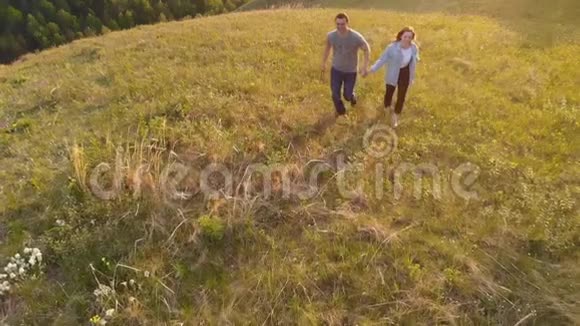 一对年轻夫妇在日落时愉快地穿过一片山地草地的航拍镜头视频的预览图