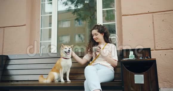 快乐的年轻女子在户外咖啡馆喝茶抚摸可爱的石坝小狗视频的预览图