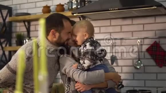 大胡子的父亲穿着温暖的毛衣在厨房里和抱着妈妈的小儿子玩耍孩子想踢视频的预览图