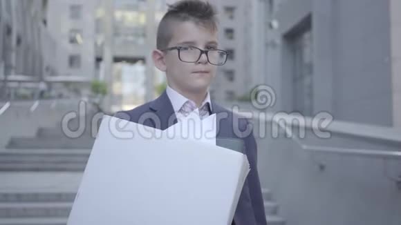 肖像可爱的小男孩穿着商务套装和眼镜拿着一个盒子在户外楼梯上放着文具儿童视频的预览图