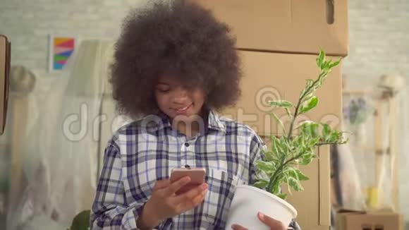 肖像非洲女人手里拿着一株植物用手机坐在盒子旁边的地板上移动视频的预览图