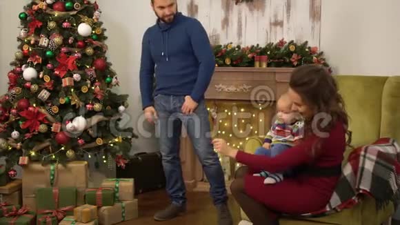 母亲父亲和小婴儿坐在装饰圣诞树附近一个人拿着小礼物盒送给视频的预览图