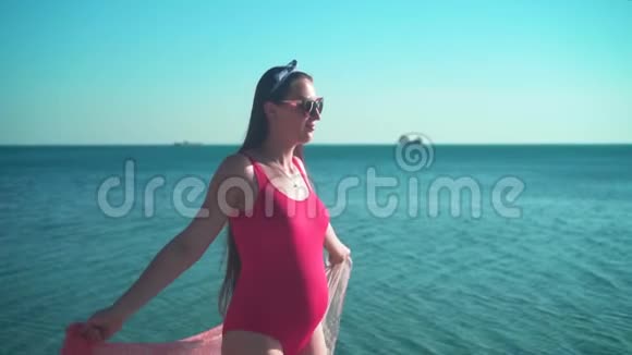 一个穿着红色一件泳衣和眼镜的怀孕女孩沿着海滨散步微笑着视频的预览图