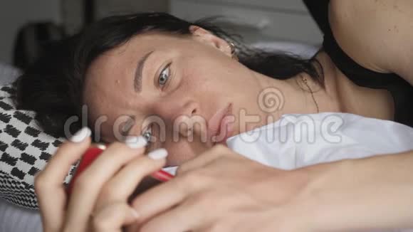 一个幸福的女人躺在她可爱的卧室里躺在白色的床上在智能手机上发短信技术互联网视频的预览图