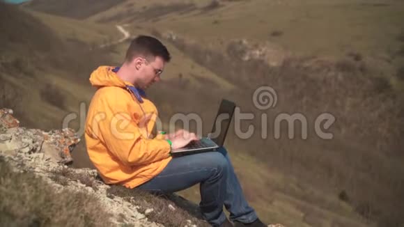 一个穿着黄色夹克蓝色牛仔裤和眼镜的年轻人坐在山上为笔记本电脑工作紧张发誓视频的预览图