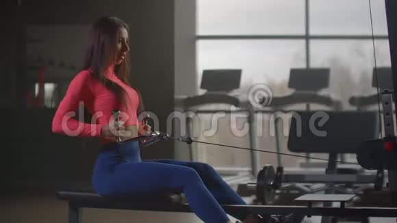 一个坐着的女人在健身房的大窗户和跑步机的背景下拉动模拟器的重量视频的预览图