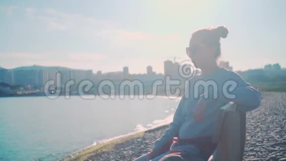 一个穿着灰色毛衣和眼镜的黑发女孩坐在海边的长凳上天空背景大海城市景观视频的预览图