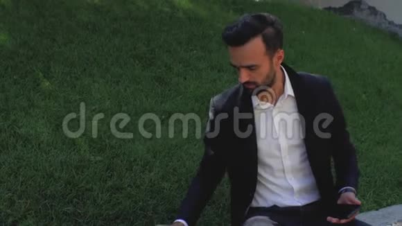 穿着经典西装留着胡子的年轻人坐在草地上的公园里喝着一次性杯子里的咖啡4k4k视频的预览图