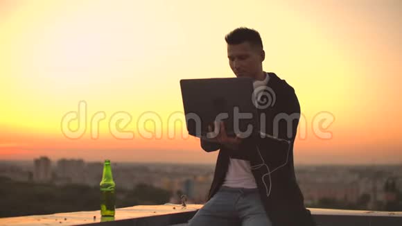 一个年轻人坐在屋顶边上拿着笔记本电脑和啤酒在笔记本电脑上打字视频的预览图