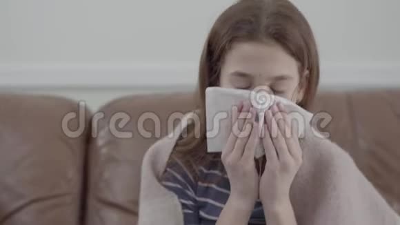 特写一幅少女的肖像裹着毯子用餐巾纸吹着鼻子坐在家里的皮革沙发上视频的预览图