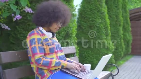 非洲裔美国妇女发型是非洲式的她用笔记本电脑坐在街边的长凳上看着摄像机视频的预览图