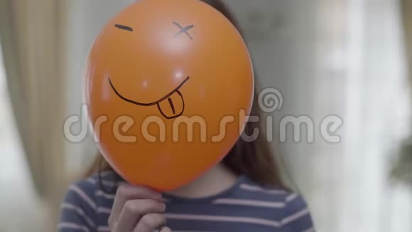 一个有趣的女孩拿着带有表情符号的橙色气球用眨眼的表情制作同样的表情符号视频的预览图