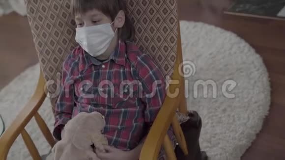 一个生病的男孩的肖像他的脸上有一个医用绷带在他的椅子上摇摆手里拿着一个软玩具在家里视频的预览图
