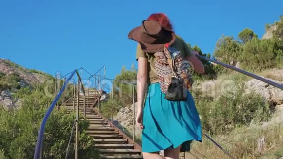 年轻的红发旅行女孩带着牛仔帽和背包爬楼梯在山区视频的预览图