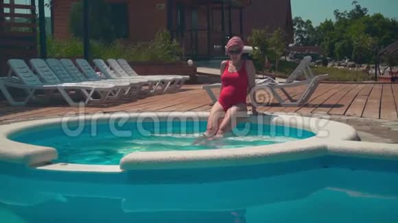 怀孕的女孩穿着红色的一件泳衣和眼镜坐在游泳池附近双腿伸进水里视频的预览图