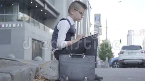 可爱的衣冠楚楚的男孩戴着眼镜坐在街上做着笔记本电脑的特写严肃的孩子像成年人一样视频的预览图