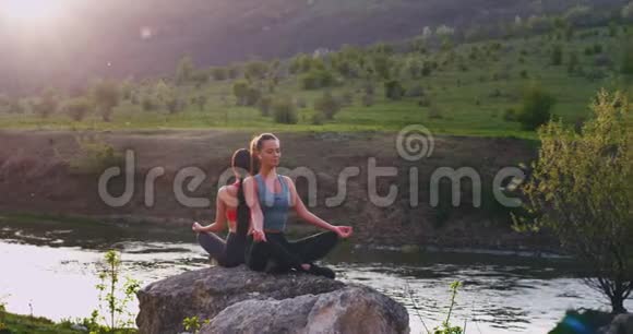 在美丽的自然景观中湖和绿野两位女士在一块大石头上做冥想姿势她们感觉到视频的预览图