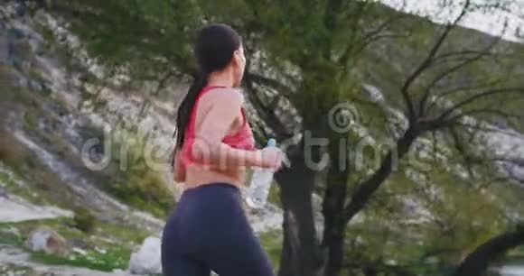 这位运动的女士在神奇的山路上奔跑一边拿着一瓶水一边听音乐视频的预览图