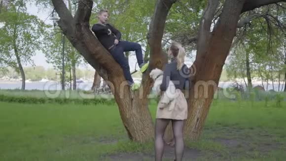 年轻的金发母亲用手机为她的小儿子在树上拍照视频的预览图