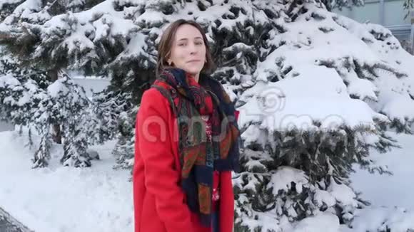 一个年轻的微笑着的黑发女孩穿着红色外套戴着围巾转过身把手放在冬天的森林景观背景上视频的预览图