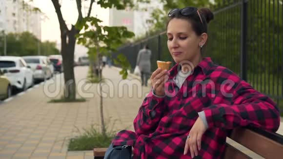 一个女孩坐在街头公园的长凳上吃冰淇淋四处闲逛那个女孩穿着一件黑色和红色的衬衫视频的预览图