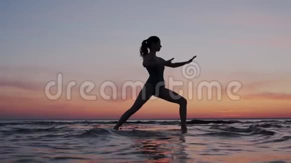 年轻的运动和柔韧的女人练习瑜伽在日落的天空中在海滩上做战士的姿势苗条和合身的剪影视频的预览图