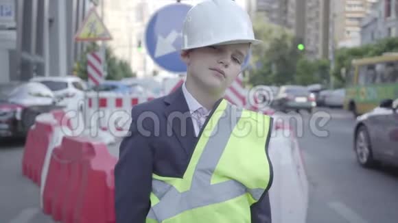 疲惫的小男孩头上戴着建筑头盔穿着制服在镜头里看着拇指建筑师概念视频的预览图