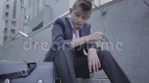 悲伤的衣冠楚楚的男孩坐在街上的楼梯上靠近他的旧钱包3那个男孩摘下眼镜感到很累视频的预览图