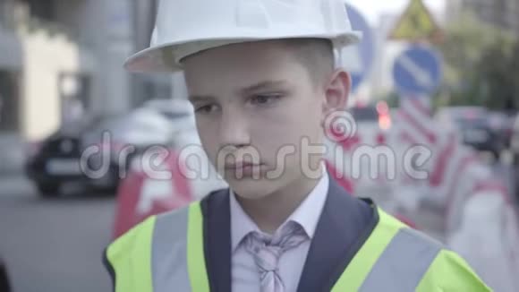 一幅悲伤疲惫的小男孩头上戴着建筑头盔的肖像制服朝远处看建筑师概念视频的预览图