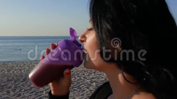 日出时一个漂亮的黑发女孩在海洋背景上喝着运动瓶中的蛋白质奶昔视频的预览图