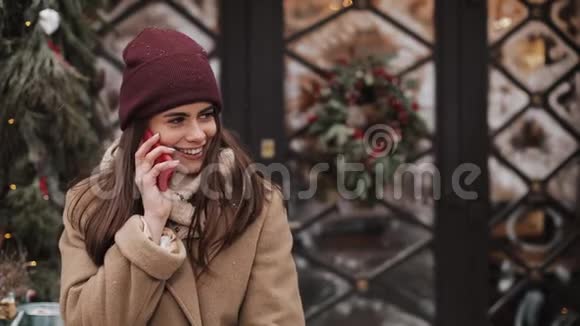 年轻的微笑迷人的女孩戴着冬帽和外套站在外面打电话看着高兴的样子视频的预览图
