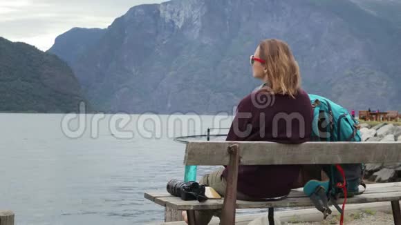 年轻的女旅行者坐在长凳上看峡湾休息视频的预览图