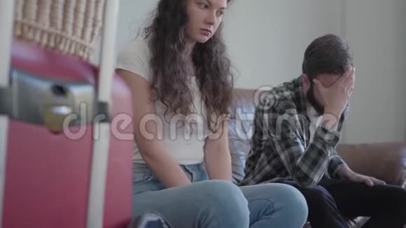 年轻夫妇在旅行前紧张地坐在家里的皮革沙发上提着旅行篮的手提箱站在附近视频的预览图