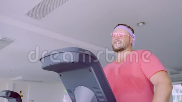 穿着粉红色T恤和粉红色眼镜的贪玩胖子在健身房里从事健身活动视频的预览图