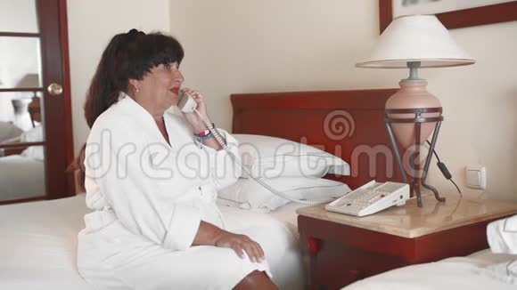 一位快乐微笑的老太太穿着白色浴袍坐在酒店的床上打电话使用的概念视频的预览图