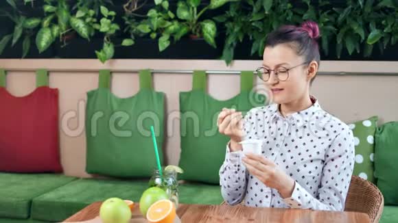 微笑着休闲的年轻女孩节食吃着新鲜的酸奶坐在咖啡馆中档的桌子上视频的预览图