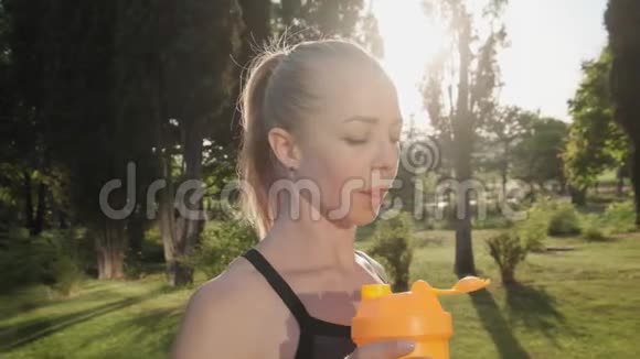 快乐运动的金发女孩在夏季慢动作的公园里晨练时喝黄色摇床上的水视频的预览图