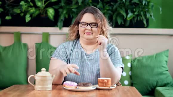 快乐丰满的女人咬甜甜圈与五颜六色的糖霜享受甜点在咖啡馆中镜头视频的预览图