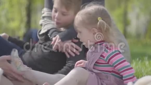 母亲在公园里系着可爱的金发小女儿的鞋而其他母亲则在坐着拥抱她的小儿子视频的预览图