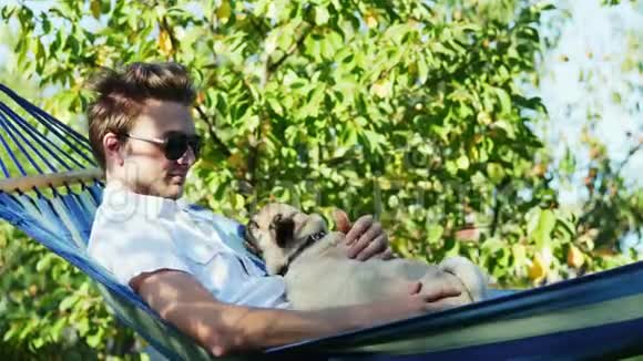戴墨镜的迷人男子和一只小狗在吊床上休息花园里平静而日落视频的预览图