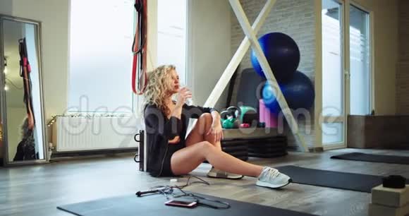 口渴的女士经过一次艰苦的健身房锻炼后她坐在地板上喝点水和轮胎度过了一段轻松的时光视频的预览图